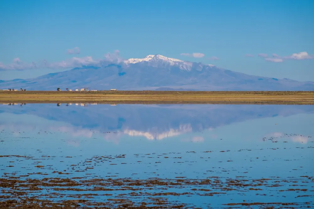 Una laguna poco profunda refleja un cerro nevado bajo el cielo azul, con una llanura amarilla en el medio
