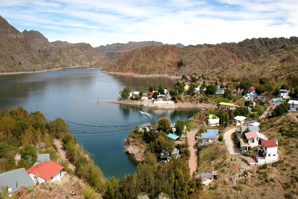 Casas y cabañas sobre la orilla de un embalse entre cerros rojizos, con una lancha que deja su estela en el agua