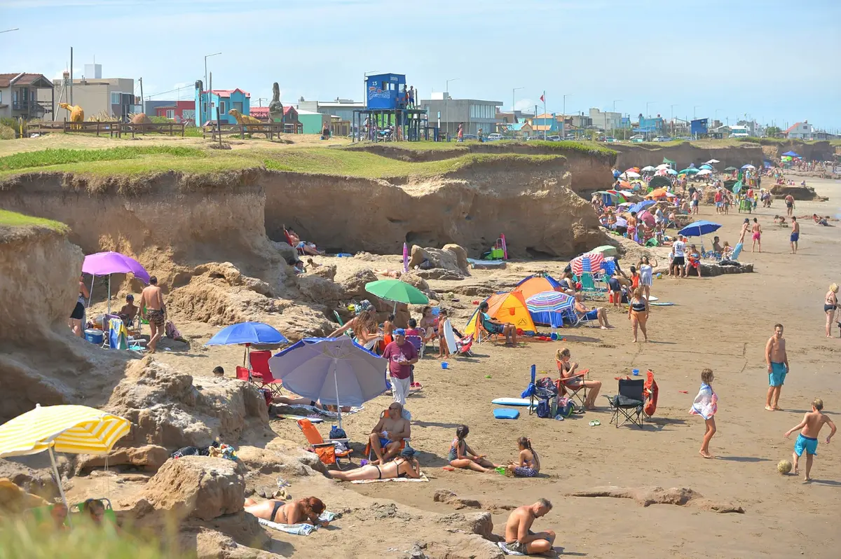 Playa con familias y sombrillas al pie de barrancas de tierra, con esculturas de dinosaurios y casas de colores arriba