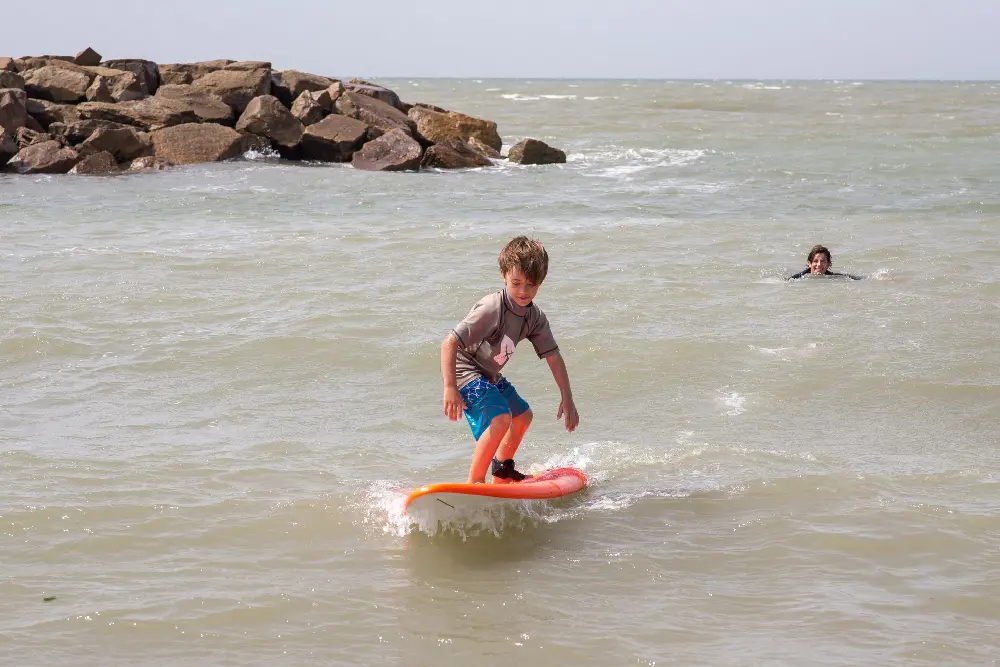 Un chico de pie sobre una tabla de surf naranja en el agua, junto a un espigón de piedras, con una mujer nadando detrás