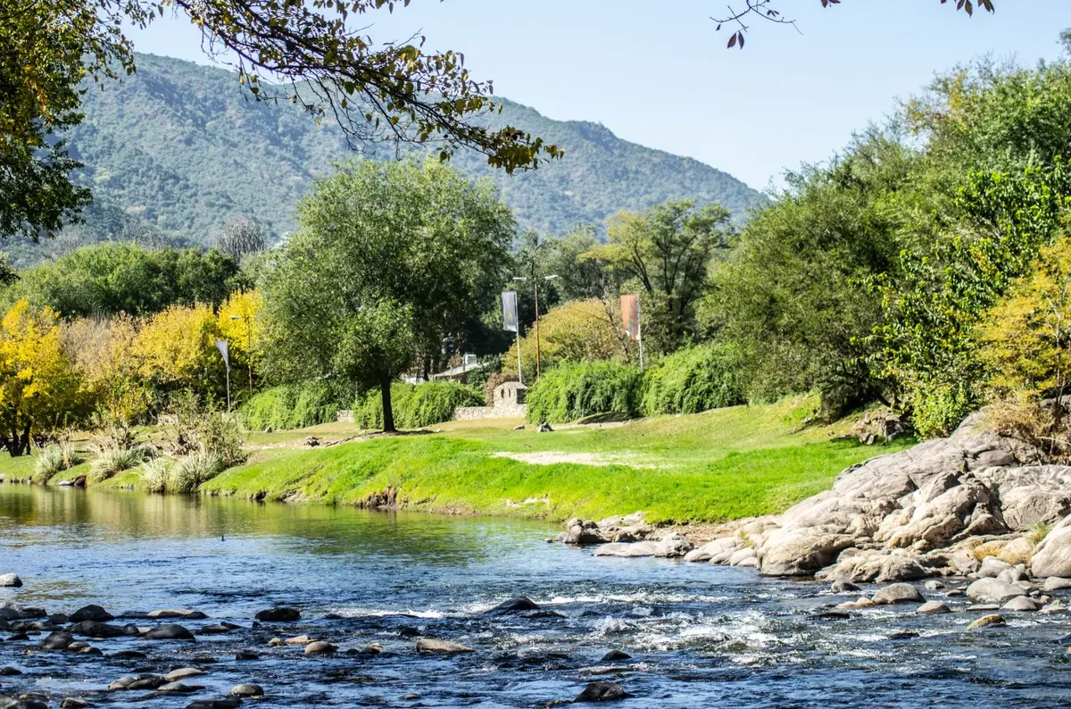 El río Santa Rosa corriendo entre piedras, con una costanera de césped y árboles y las sierras de fondo