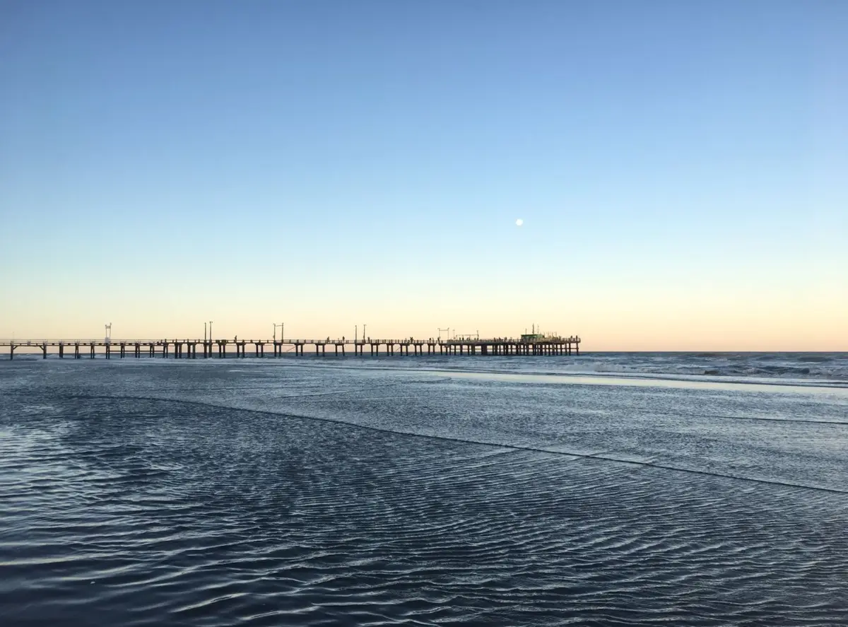 Muelle largo recortado contra el cielo del atardecer, con la luna sobre el horizonte y el agua de la orilla en primer plano