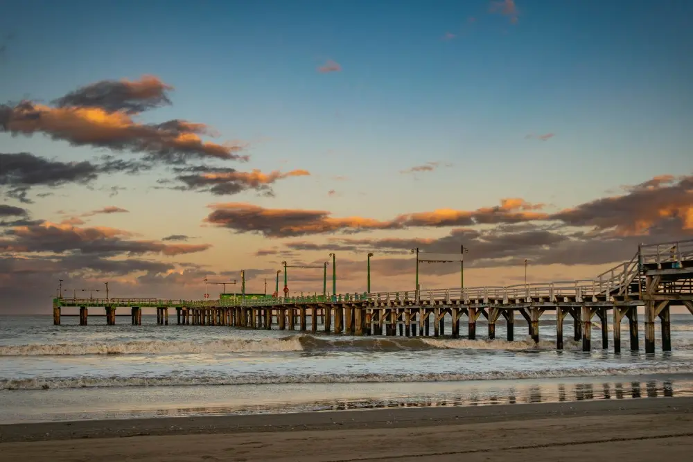 Muelle de madera con barandas verdes que entra en el mar al atardecer, con nubes anaranjadas y olas rompiendo sobre la orilla