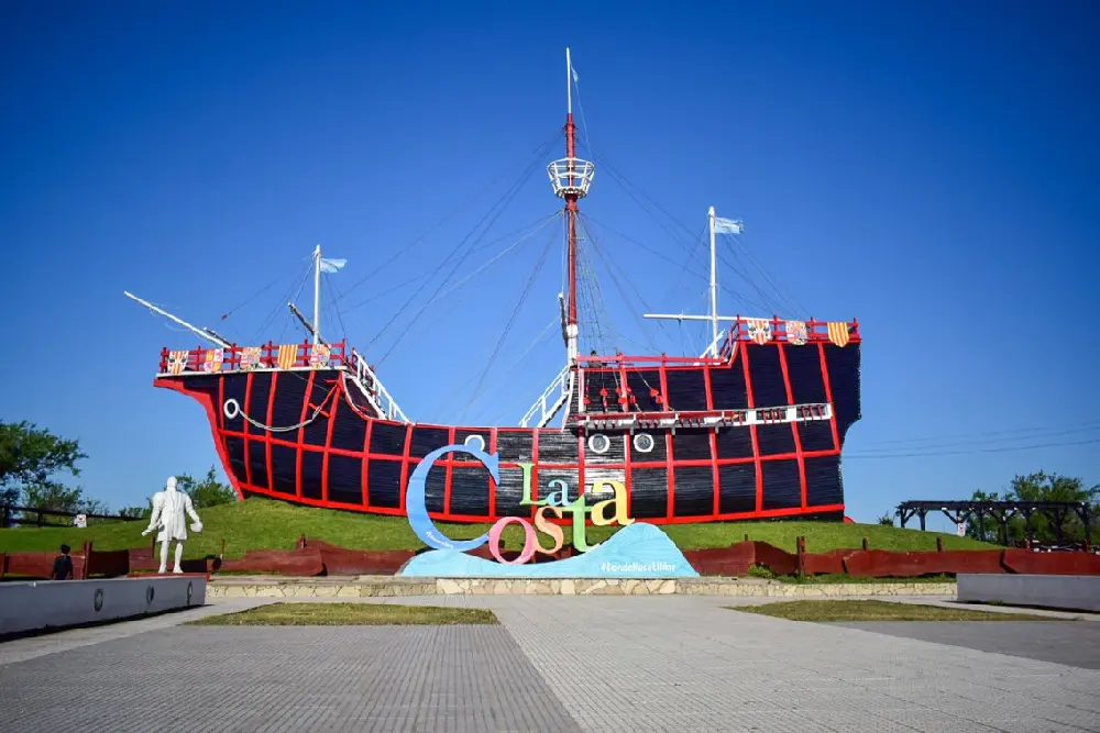 Réplica de una carabela con el casco negro y rojo sobre un montículo de césped, con una estatua blanca al costado y un cartel de colores que dice La Costa