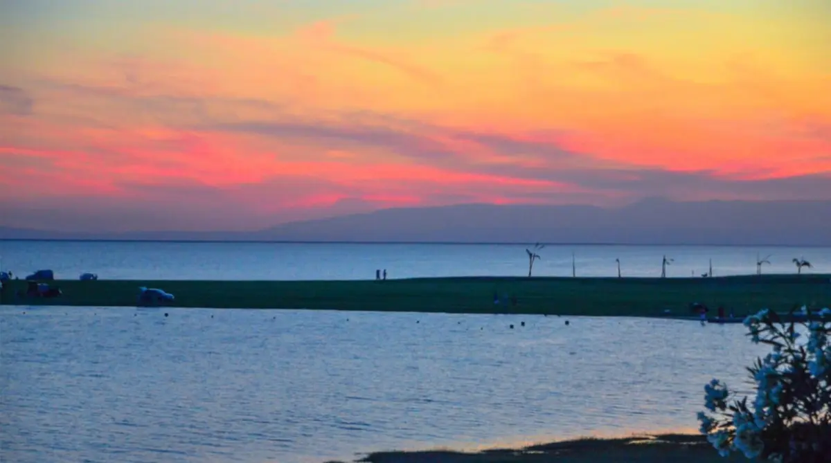 Atardecer rosado y naranja sobre un gran espejo de agua, con una franja de costa verde y sierras en el horizonte