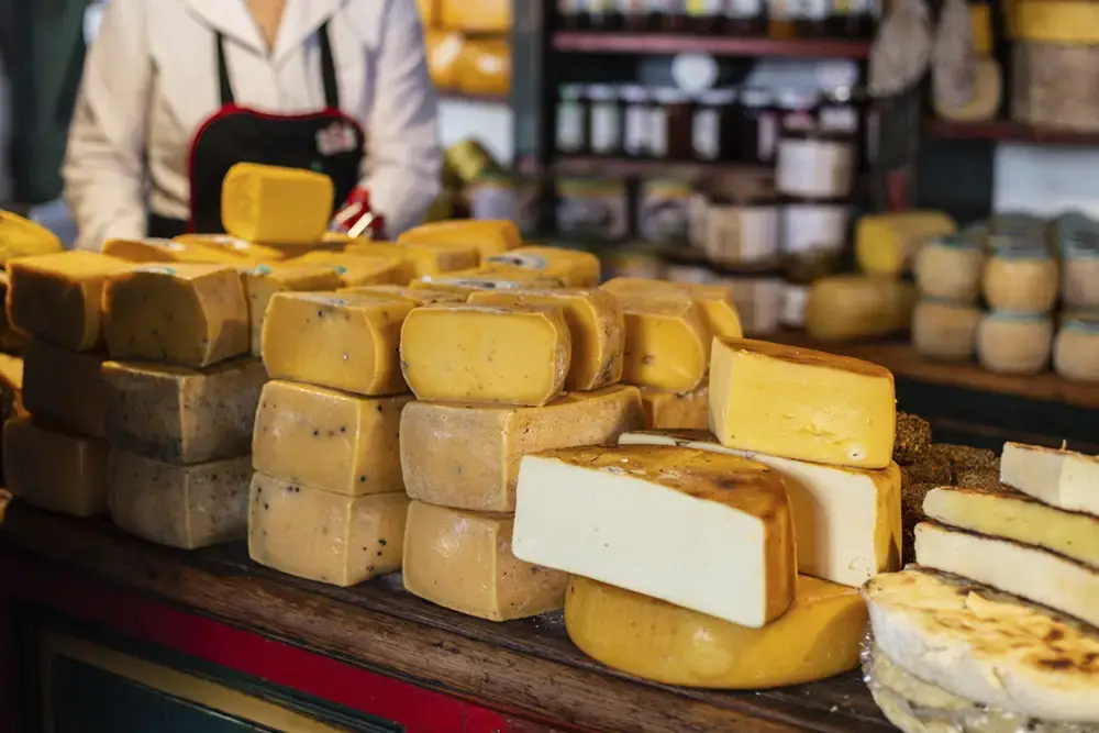 Hormas de queso apiladas sobre el mostrador de madera de un almacén, con frascos de conservas en los estantes