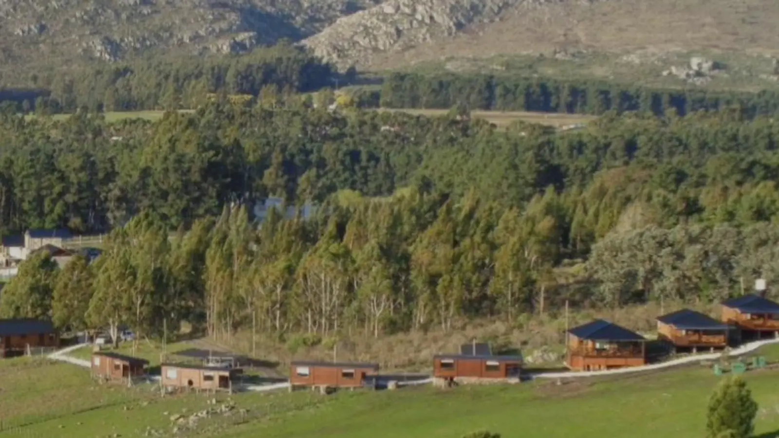 Cabañas de madera sobre una ladera verde, con un bosque de pinos y las sierras de piedra de Tandil al fondo