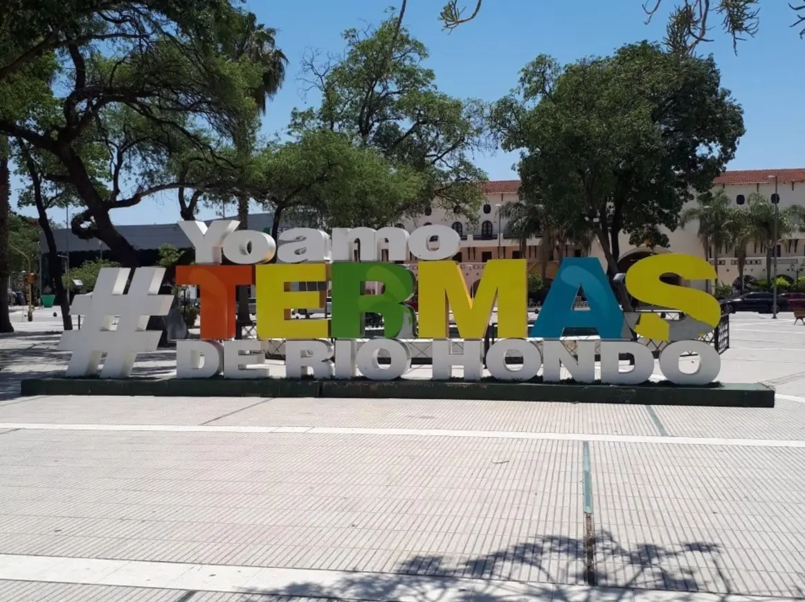 Letras de colores que dicen Yo amo Termas de Río Hondo en una plaza, con árboles y un edificio blanco detrás