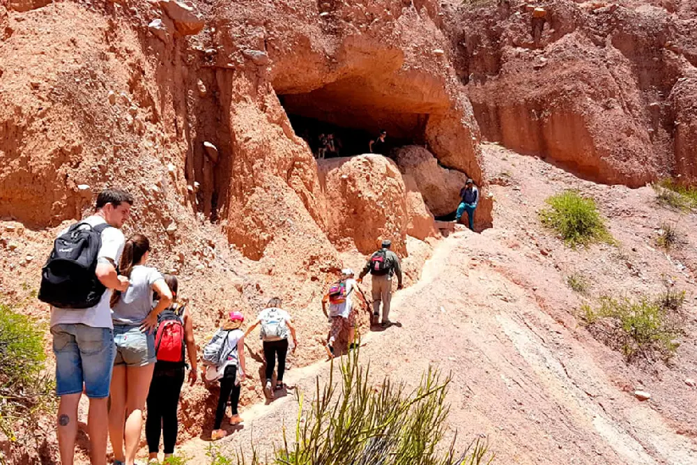 Grupo de caminantes con mochilas subiendo por un sendero de tierra rojiza hacia la boca de una cueva