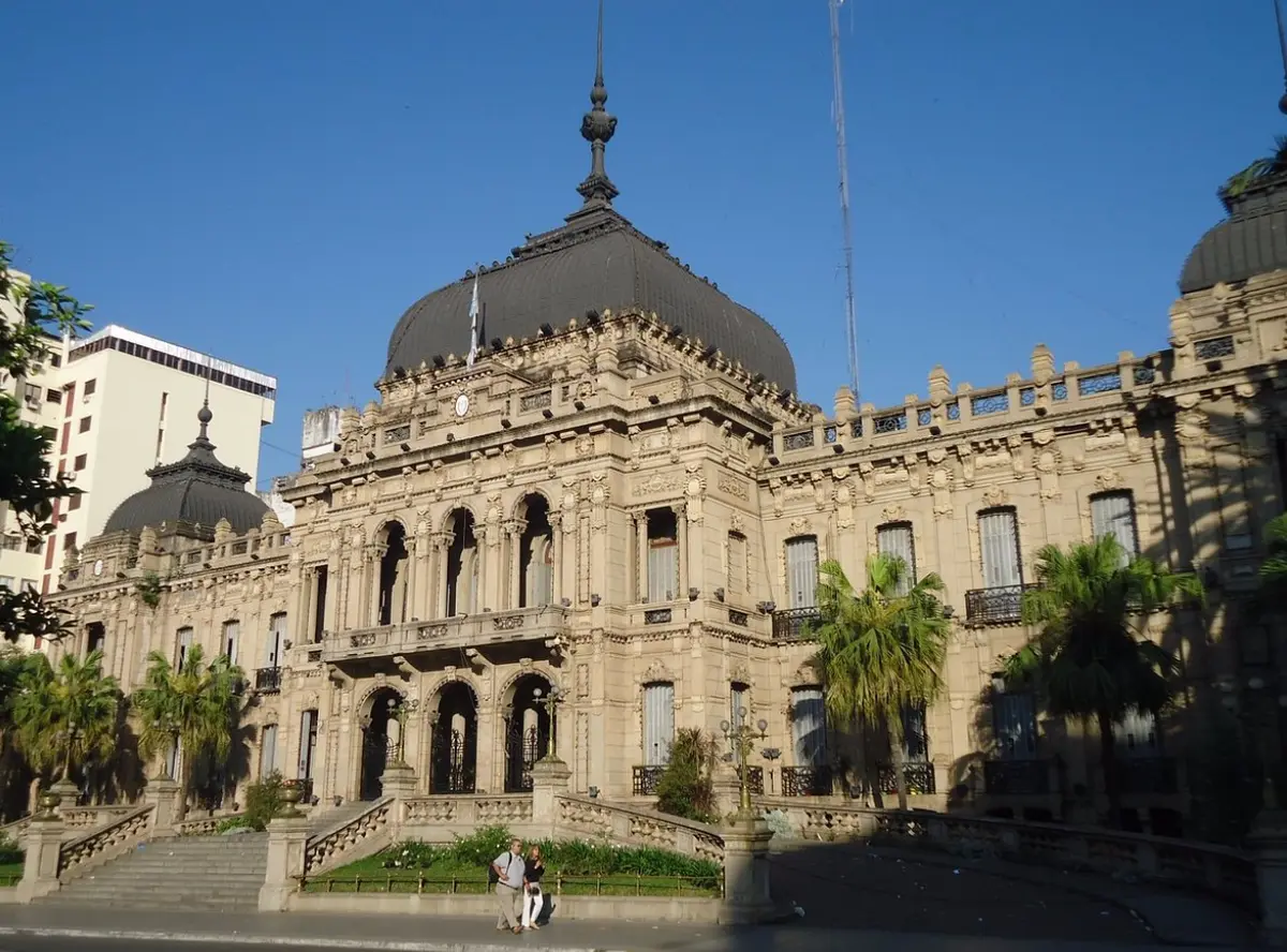Fachada de la Casa de Gobierno de Tucumán con su cúpula central, escalinata, palmeras y dos personas caminando