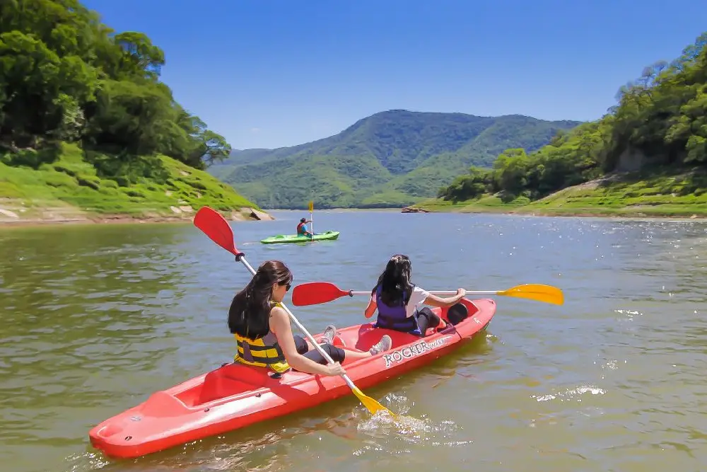 Dos chicas reman en un kayak rojo por un embalse rodeado de cerros cubiertos de selva, con otro kayak más adelante