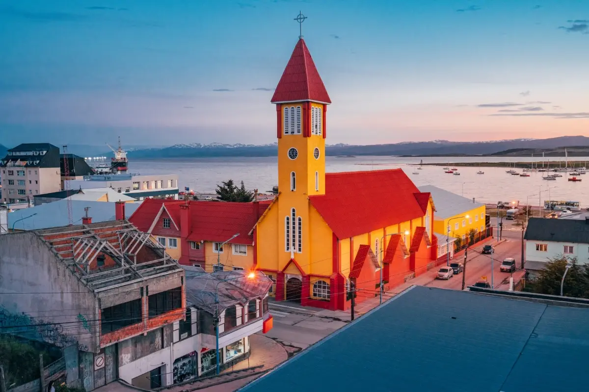 Iglesia de paredes amarillas y techo rojo en el centro de Ushuaia al atardecer, con la bahía y los barcos de fondo