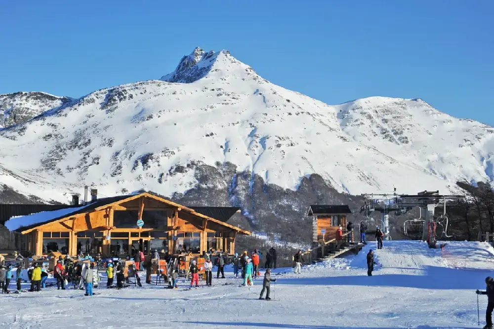 Esquiadores frente a un refugio de madera al pie de un cerro completamente nevado, junto a la base de una aerosilla