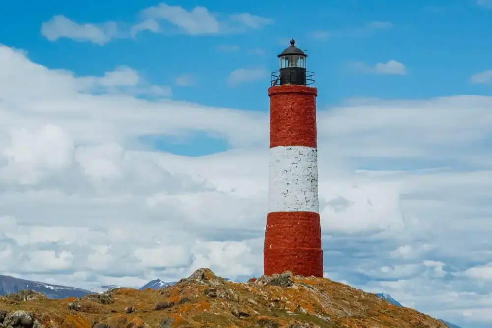 Faro de franjas rojas y blancas sobre un islote rocoso con líquenes anaranjados, con nubes blancas y montañas al fondo