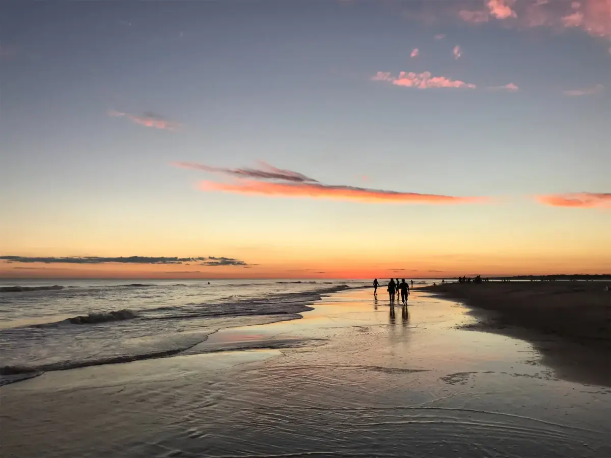 Personas caminando por la orilla mojada al atardecer, con el cielo naranja y rosado reflejado en la arena