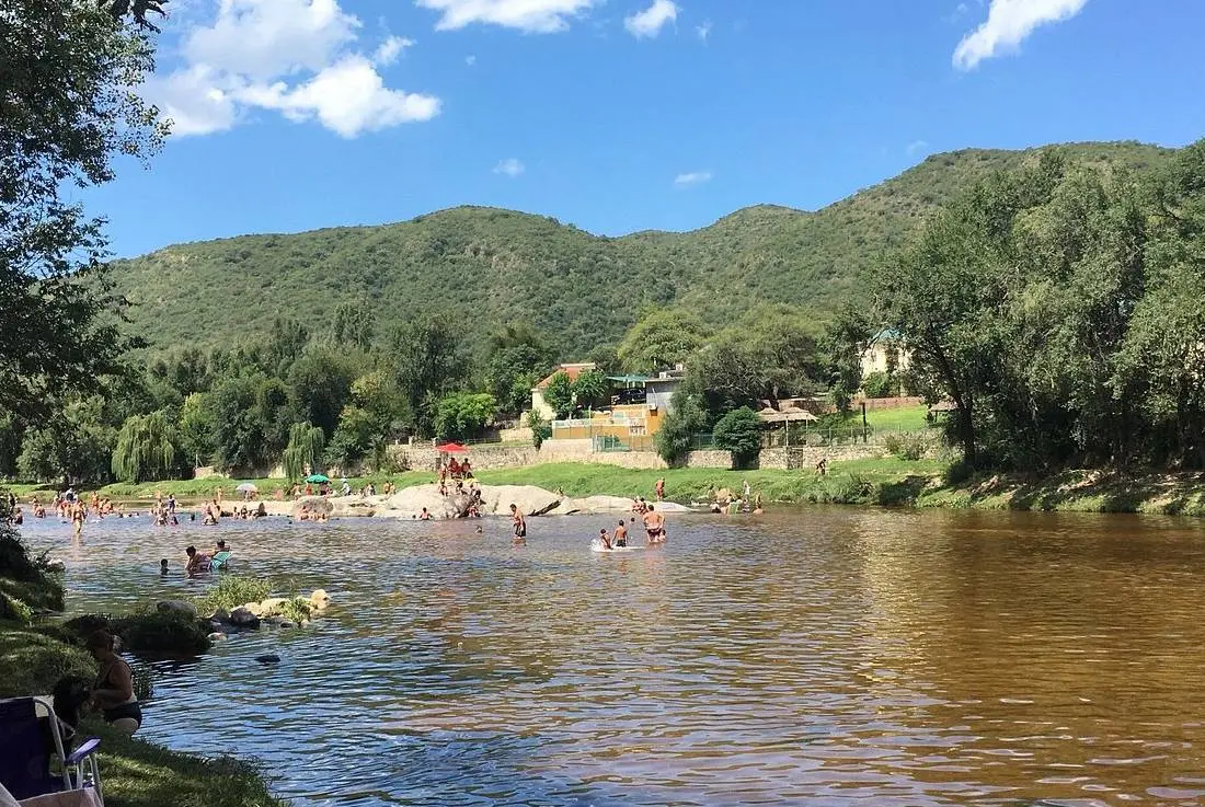 Gente bañándose en un balneario de río con piedras, sauces y las sierras verdes de fondo