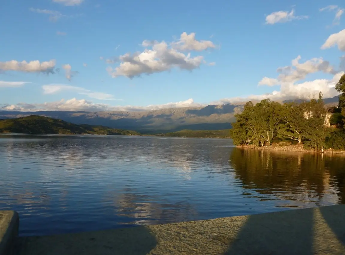 El lago del dique bajo un cielo con nubes, con una punta arbolada a la derecha y el cordón de sierras al fondo