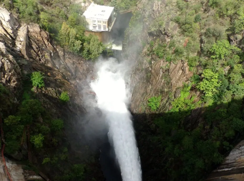 Chorro de agua blanca que cae con fuerza en un cañón de paredes de roca y vegetación, bajo un edificio en lo alto