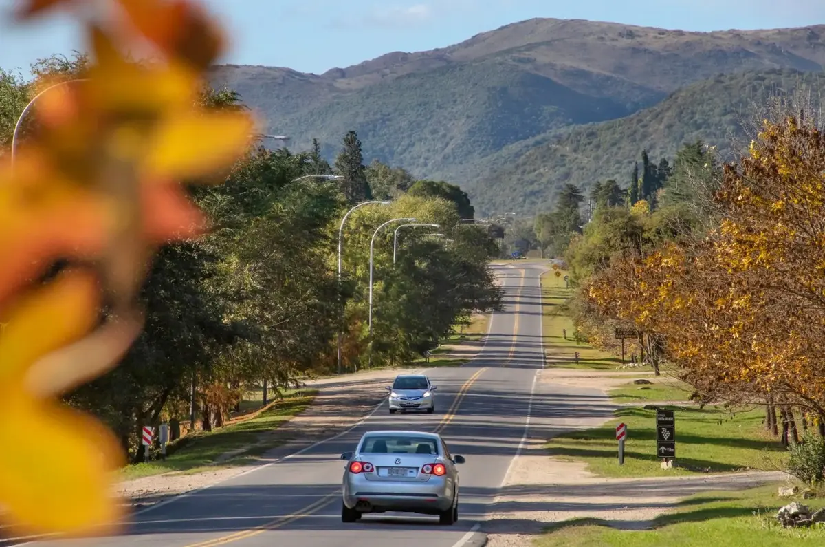 Ruta arbolada de Calamuchita en otoño, con autos y las sierras de fondo