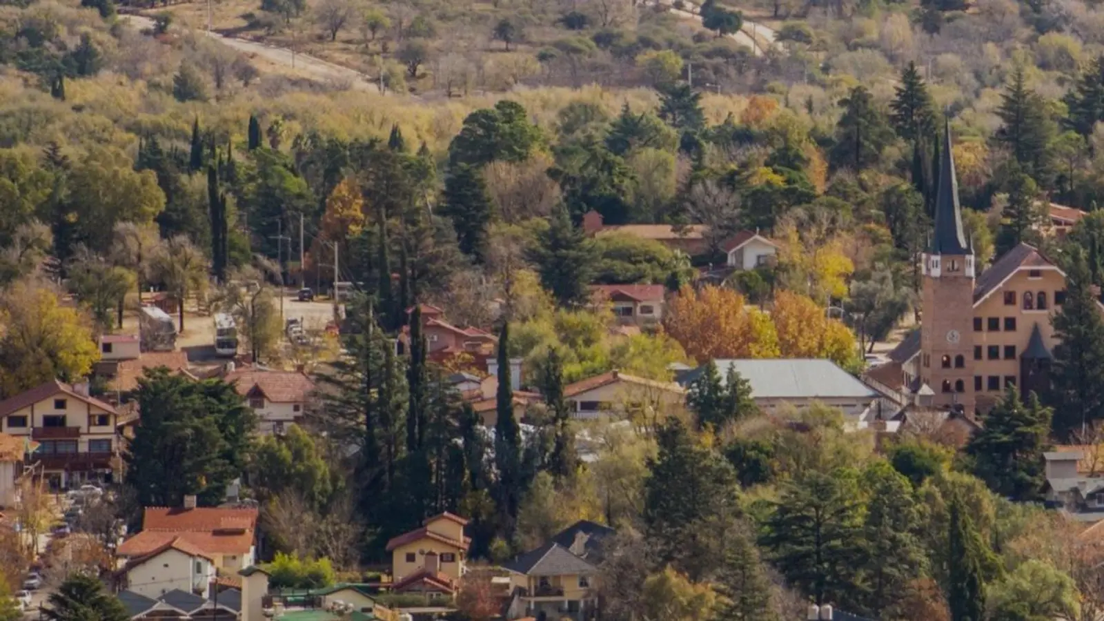 Vista de Villa General Belgrano en otoño, con casas de techos rojos entre pinos y árboles dorados y una torre de iglesia