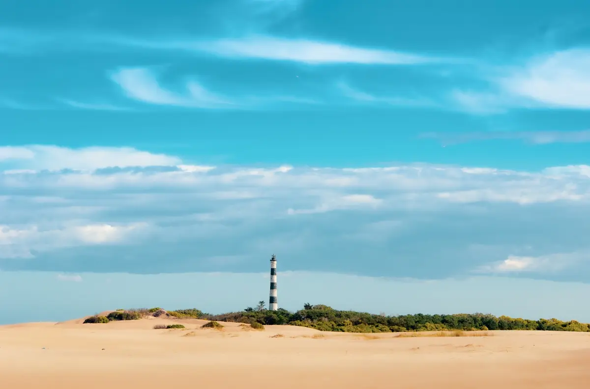 El Faro Querandí, con sus franjas blancas y negras, asomando sobre los médanos y el monte bajo, bajo un cielo con nubes