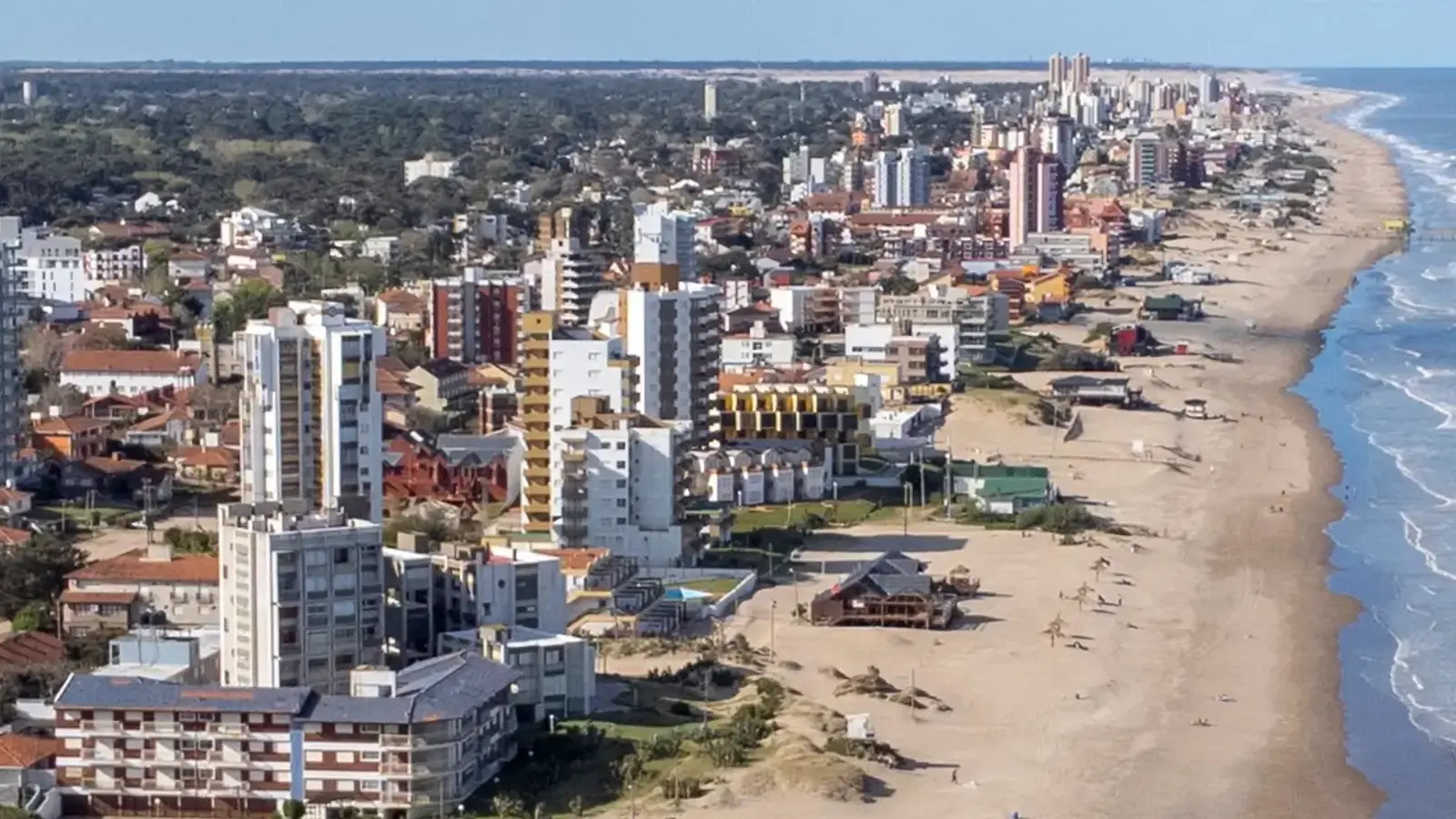 Vista aérea de la costa de Villa Gesell, con edificios frente a una playa ancha, médanos, paradores y el bosque detrás