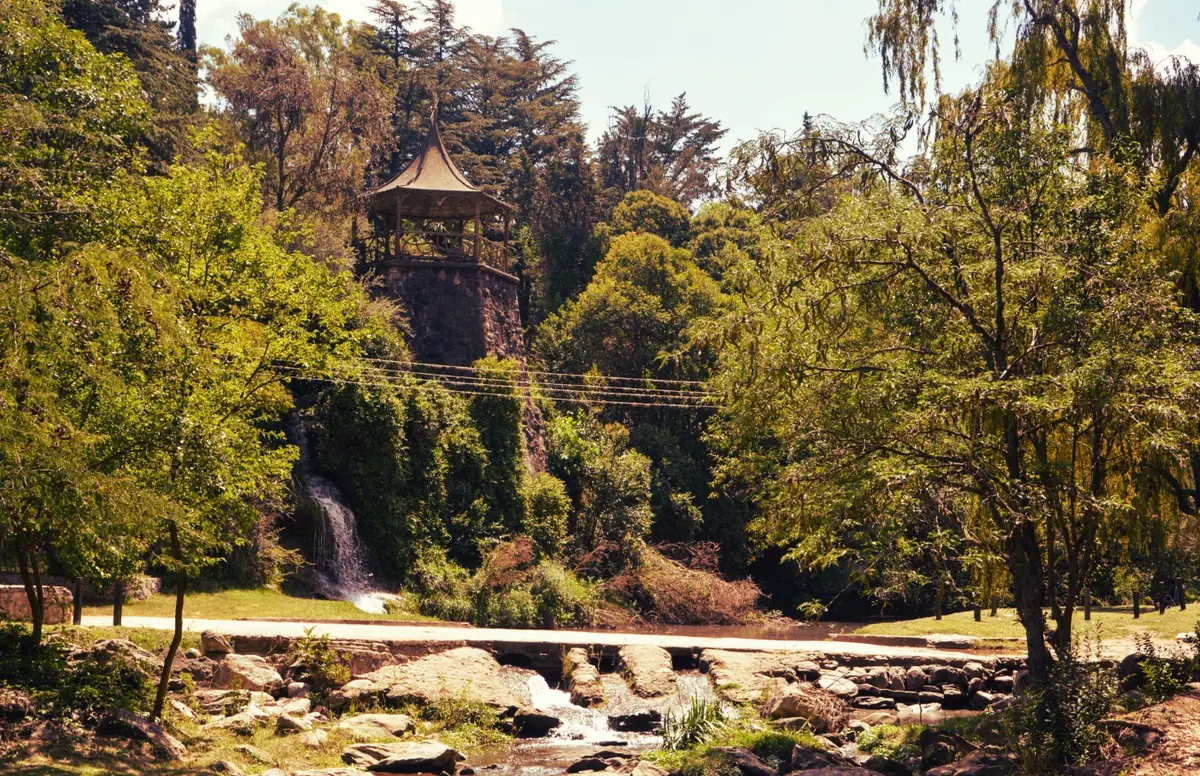 Torre de piedra con un techo de madera en punta entre árboles altos, junto a una pequeña cascada y un río con rocas