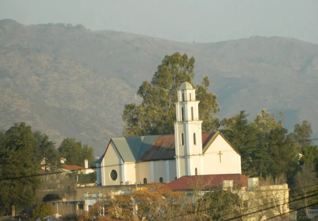 Iglesia blanca con campanario alto y techo de chapa entre árboles, con las sierras de fondo