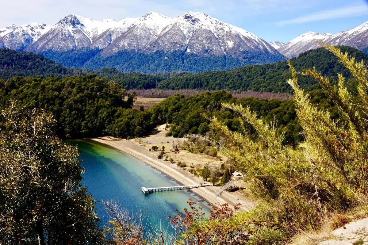 Una playa de arena con un muelle de madera sobre un lago turquesa, rodeada de bosque y con montañas nevadas de fondo