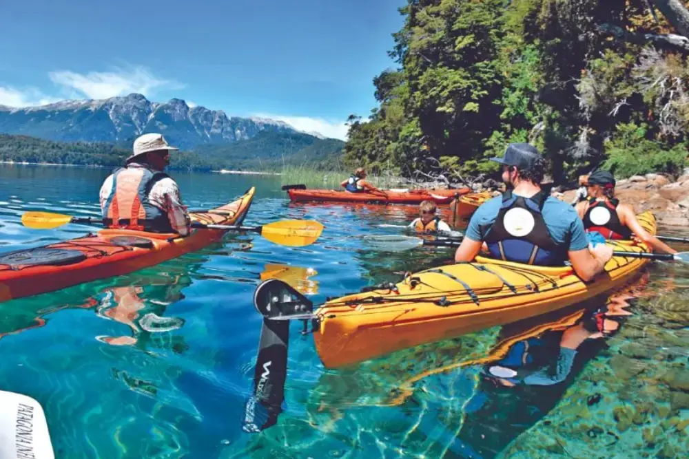 Varias personas en kayaks de colores sobre un lago de agua transparente, junto a una costa de bosque y con montañas al fondo