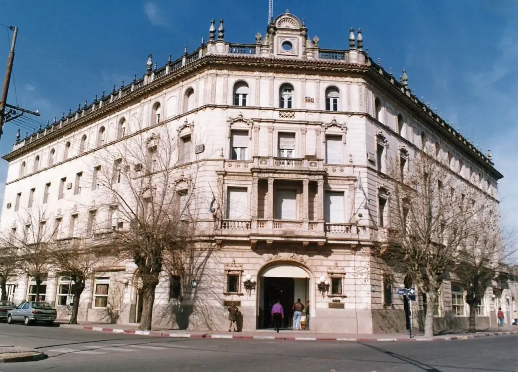 Edificio histórico de cuatro pisos en una esquina, con fachada clara, balcones y una cornisa ornamentada, bajo el cielo azul