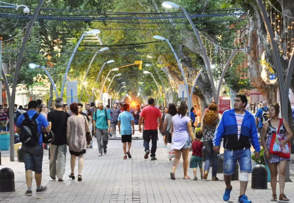 Una calle peatonal arbolada con faroles curvos y luces colgantes, llena de gente caminando