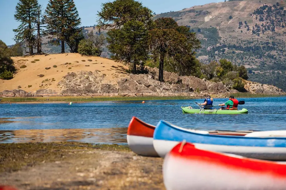 Dos personas reman un kayak verde en un lago, con canoas de colores en la orilla y araucarias sobre una loma