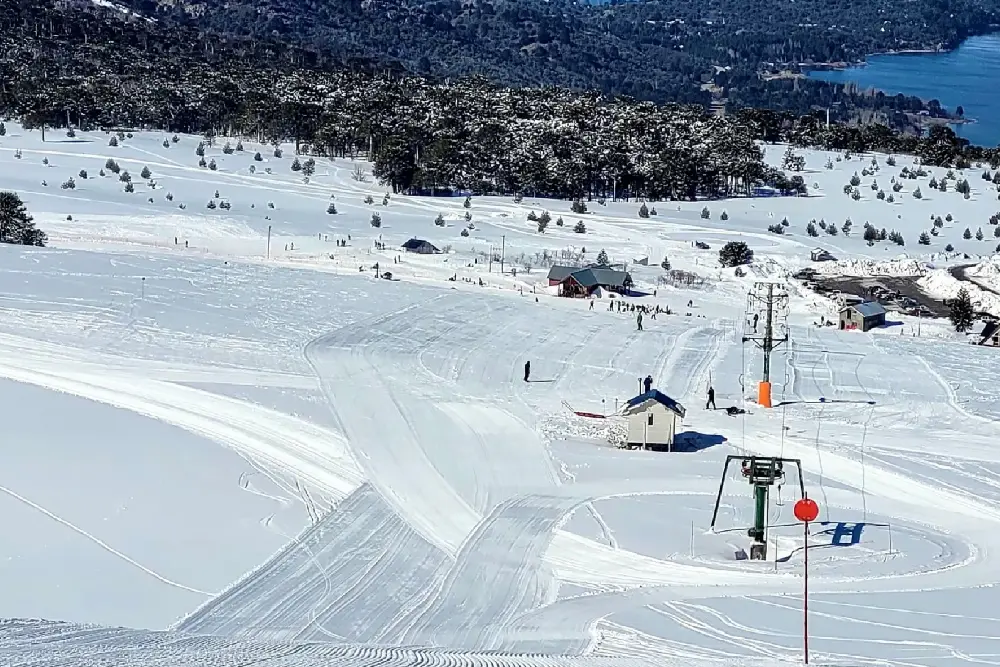 Pistas de esquí nevadas con medios de elevación y una casita de madera, con bosque y un lago al fondo