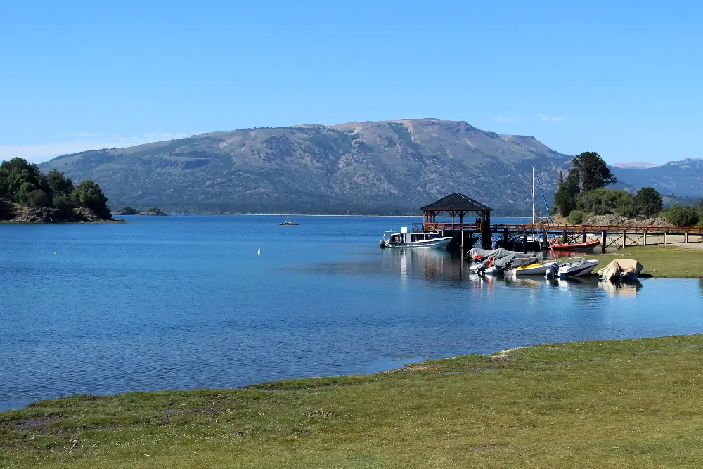 Muelle de madera con un quincho y lanchas en un lago calmo, con un cerro de cima chata al fondo
