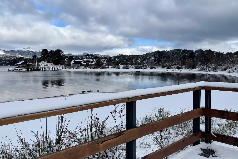 Baranda de madera cubierta de nieve frente a un lago calmo, con un muelle y casas entre árboles nevados