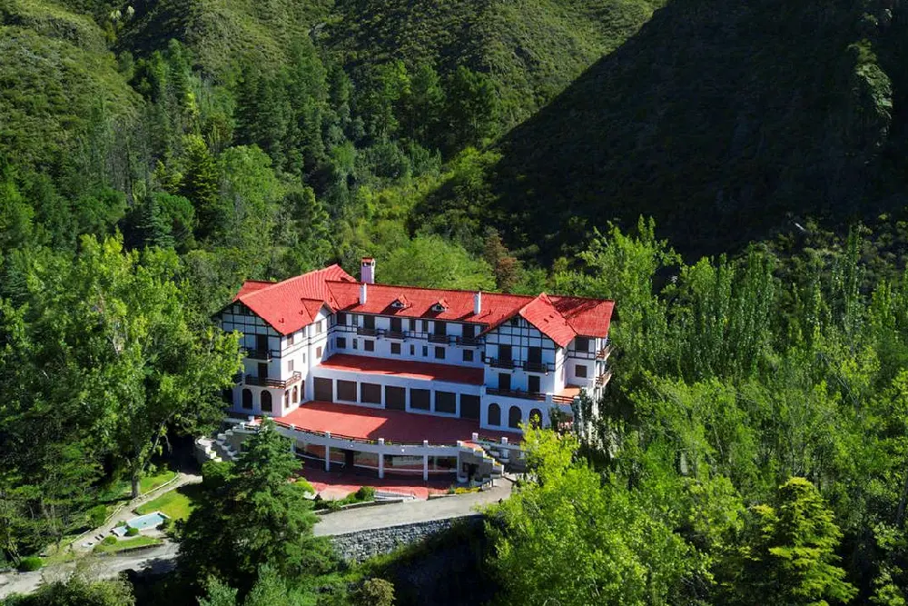 Vista desde lo alto de un gran hotel blanco de techos rojos y estilo normando, rodeado de árboles en una quebrada verde