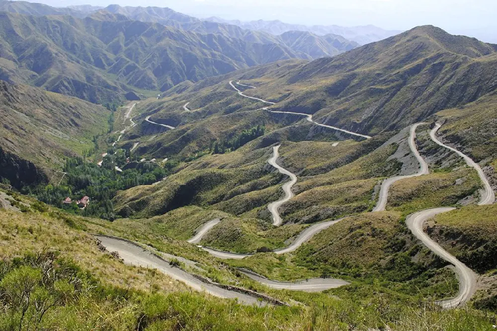 Camino de ripio que sube en curvas cerradas por laderas de montaña verdes, con un caserío entre árboles al fondo del valle